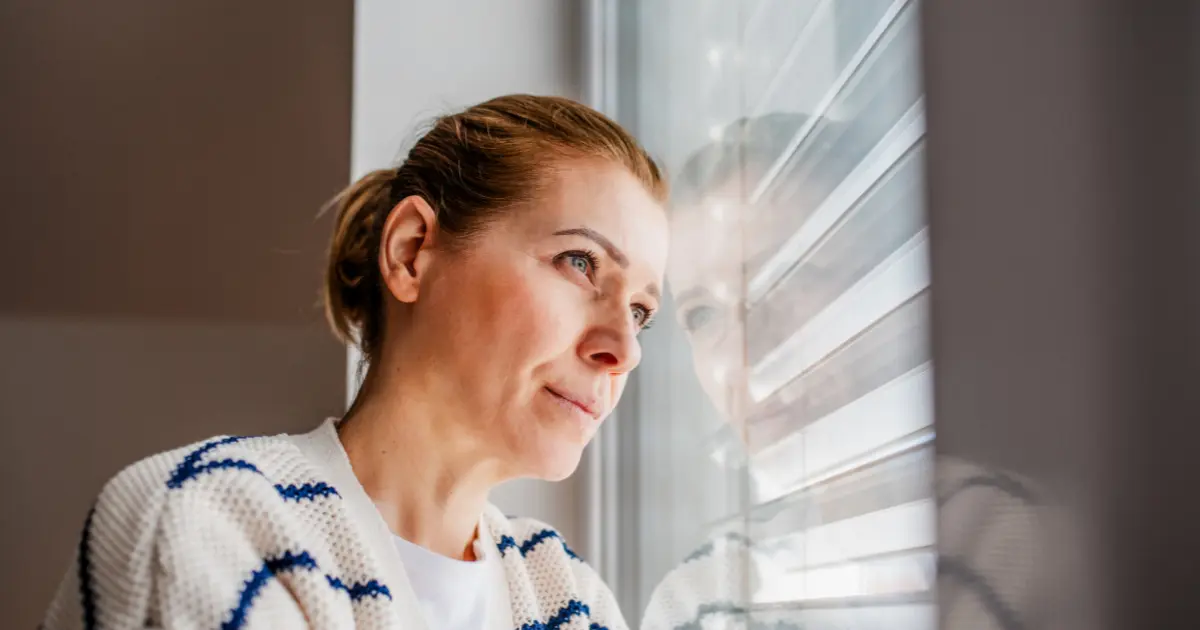 Woman in a white and navy cardigan gazing through a window with her reflection visible in the glass.